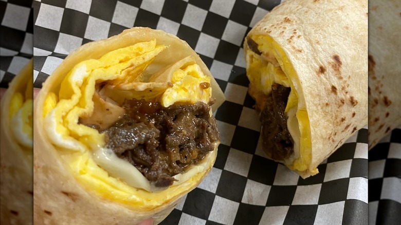 A breakfast burrito cut in half on black and white checkered food liner, with eggs, cheese, and bulgogi inside