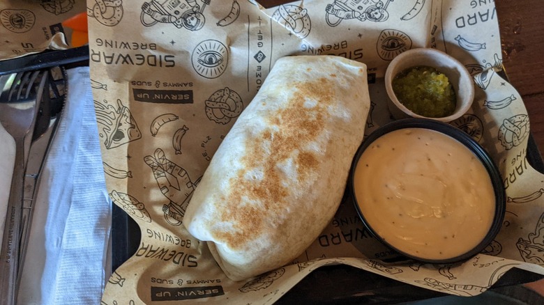 A massive burrito on a food liner with the Sideward Brewing logo on it, next to a bowl of beer cheese, and a side of guacamole