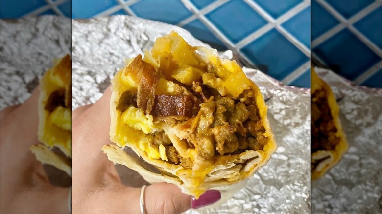 A close-up of a hand holding half a breakfast burrito that's stuffed with potatoes, eggs, and meat