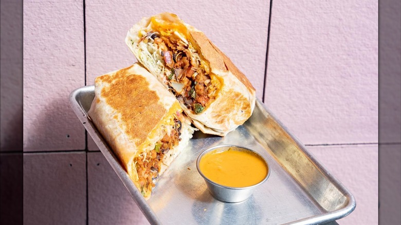 An aluminum tray against a pink wall that has a currito cut in half with a side of queso next to it .The burrito is stuffed with tater tots, beans, cabbage, pico de gallo, crema, and salsa ranchera