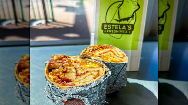A burrito cut in half stuffed with eggs, cheese, bacon, potatoes, and an entire pancake with an Estela's Fresh Mex bag in the background