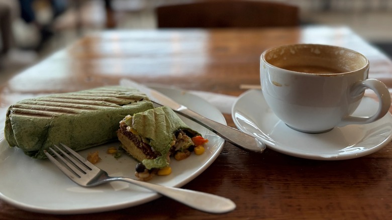 A grilled burrito on a plate that's been bitten into next to a half-drunk cup of hot coffee