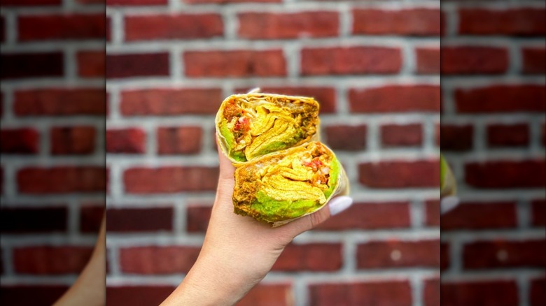 A hand holding a breakfast burrito cut in half, with scrambled eggs, avocado, hash browns, and pico de gallo against a brick wall background