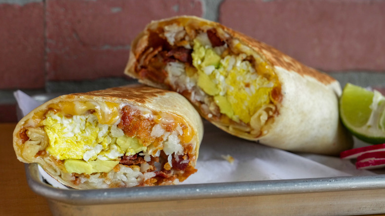 A breakfast burrito cut in half, filled with eggs, bacon, hash browns, avocado, and salsa, served on a tray with lime