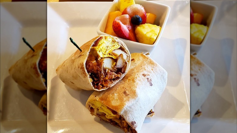 A breakfast burrito cut in half with eggs, potatoes, and meat, served with a side of fresh fruit