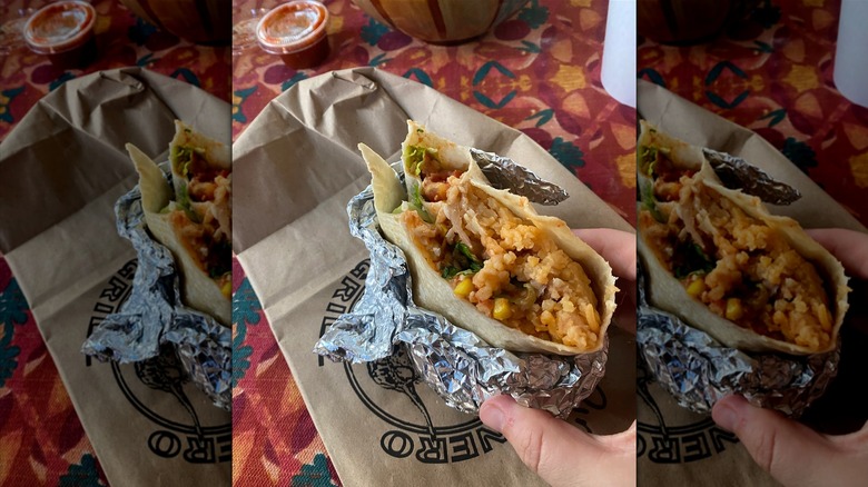 A foil-wrapped breakfast burrito filled with eggs, rice, corn, and vegetables, held over a brown paper bag with salsa on the side