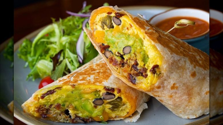 A breakfast burrito cut in half, filled with cheesy eggs, black beans, avocado, bacon, and jalapeños, served with a side salad and salsa
