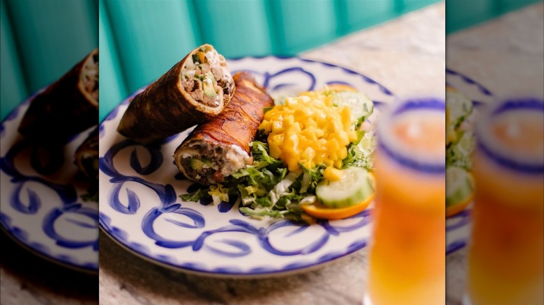 A breakfast burrito cut in half, filled with steak, beans, pico, avocado, and Oaxaca cheese, wrapped in crispy bacon and served with a side salad