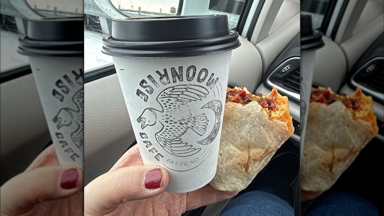 A hand holding a takeout coffee and a breakfast burrito filled with eggs, potatoes, and bacon inside a car
