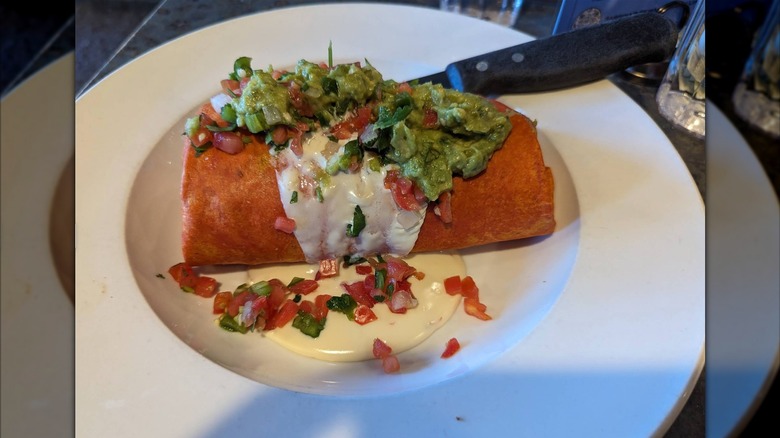 A smothered breakfast burrito topped with guacamole, pico de gallo, and sour cream on a white plate with a knife