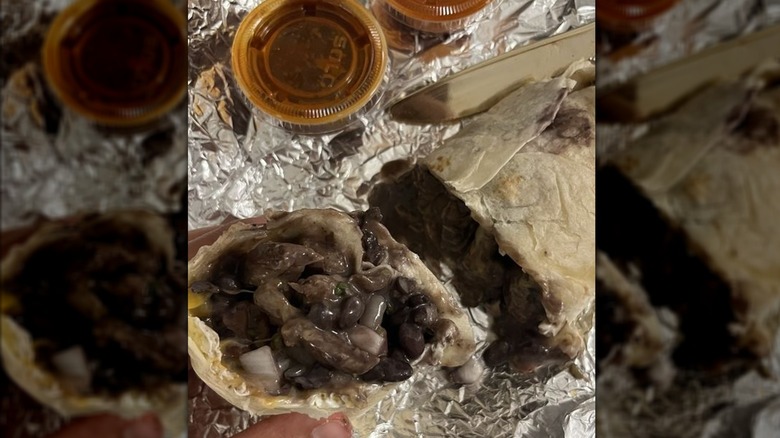 Someone holding half a breakfast burrito loaded with eggs, beans, steak, and onions with the other half in the background