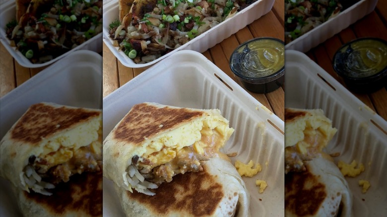 A grilled breakfast burrito, filled with potato, chorizo gravy, and eggs in a takeout container with a green sauce next to it