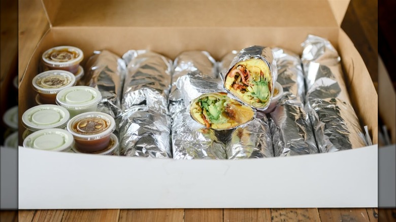 A box of foil-wrapped breakfast burritos with sides of salsa, one cut open to show eggs, bacon, and avocado
