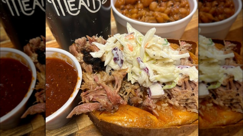 Shredded meat and coleslaw on top of sweet potato next to a bowl of baked beans, a red dip, and drink.