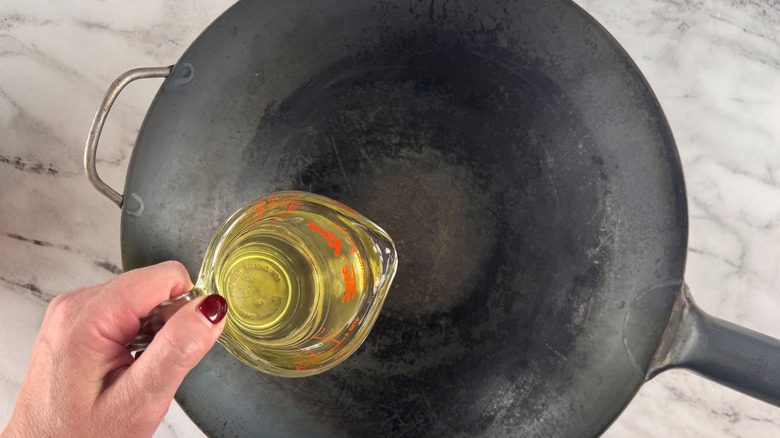 Hand holding measuring cup of oil over wok