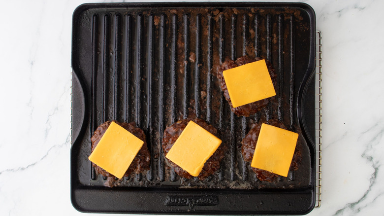Burger patties topped with cheese slices on dirty grill pan