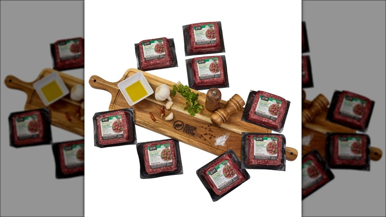Packages of ground beef surrounding a wooden cutting board with oil, herbs, garlic, and other items.