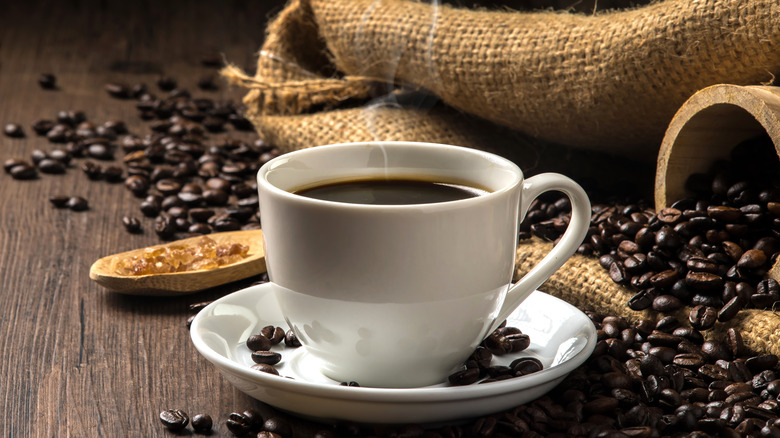 A hot cup of coffee on a wood table surrounded by coffee beans