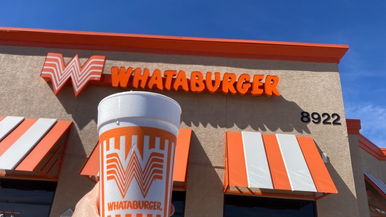 Hand holding a soft drink cup in front of Whataburger storefront during daytime