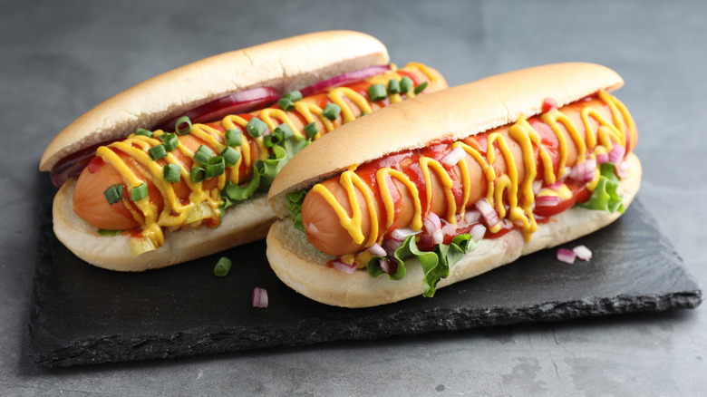 Two hotdogs on a slate board, each decorated with onions, mustard, ketchup and other accoutrements