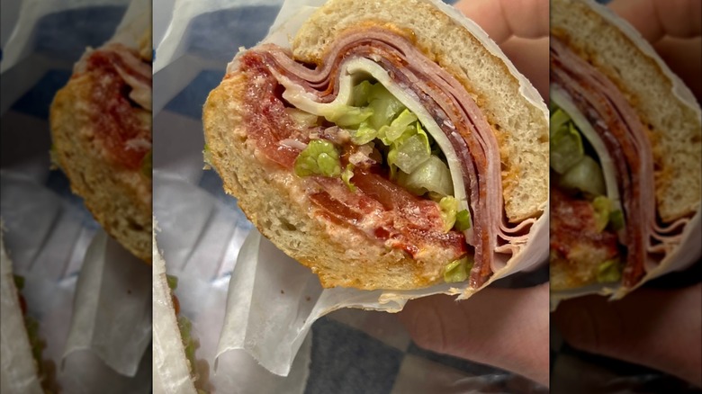 A close-up of an Italian sub showing layers of salami, ham, provolone, shredded lettuce, and dressing on a soft roll