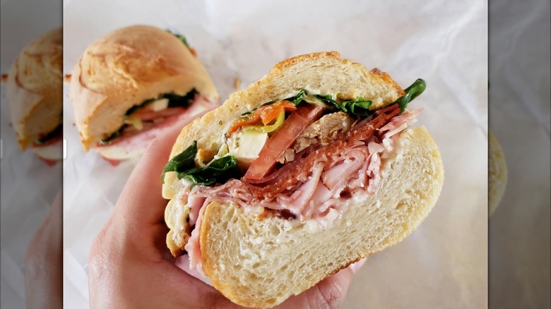 A hand holding anItalian sub, filled with ham, salami, pepperoni, provolone, greens, and a dressing