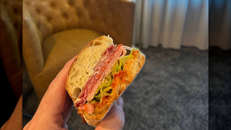 A hand holding half of an Italian sub stacked with salami, ham, shredded lettuce, and a bright orange dressing on soft bread