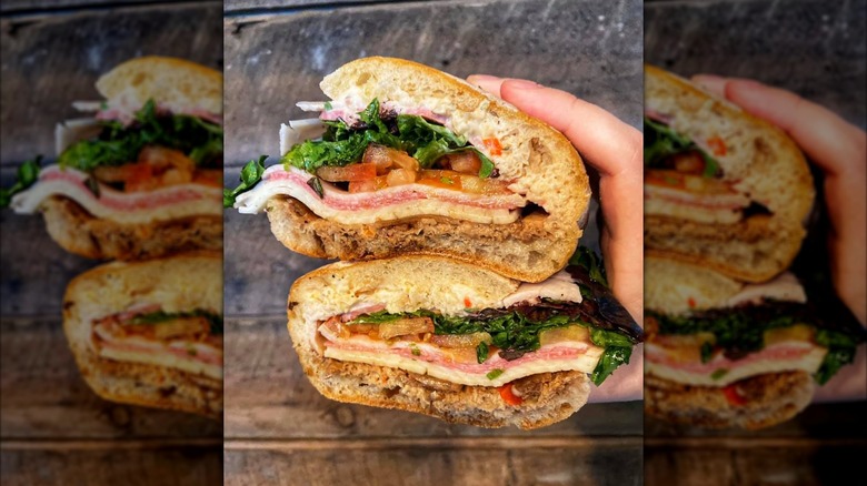 Two halves of an Italian sub stacked in hand, with layers of deli meats, cheese, tomato, and lettuce on a crusty roll