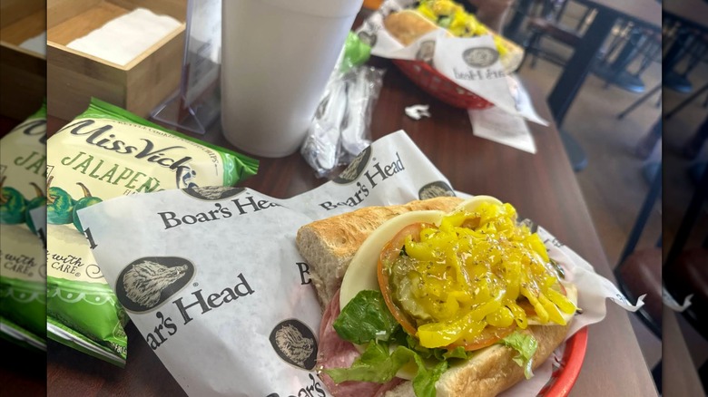 An Italian sub with deli meats, cheese, tomato, lettuce, and bright yellow banana peppers, served in a basket lined with Boar's Head paper alongside a bag of jalapeño chips and a drink