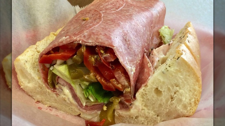 An Italian sub with layers of deli meats and provolone, lettuce, tomato, and peppers, with a slice of salami folded over the top to keep everything inside