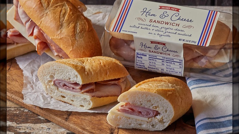 A ham and cheese sandwich sits on a cutting board with a packaged sandwich in the background.