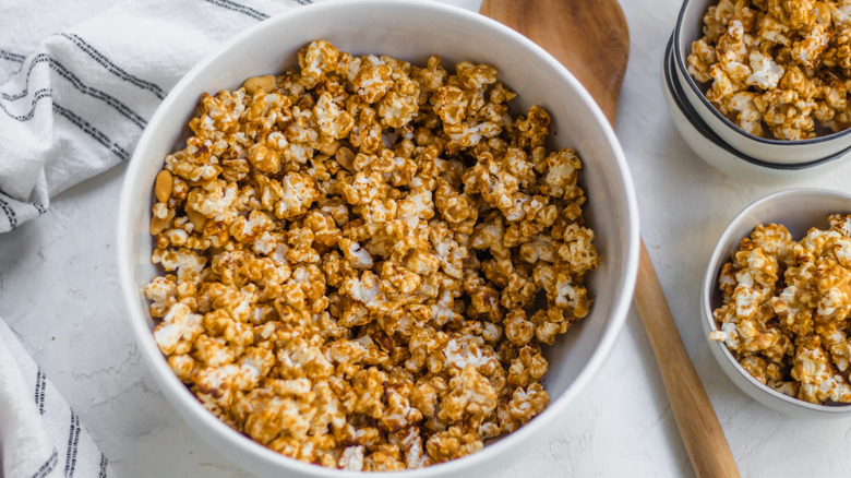 Peanut butter popcorn in white bowls on white table with kitchen towel and wooden spoon
