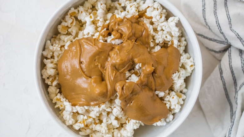 Peanut butter sauce on unflavored popcorn in white bowl atop white table with kitchen towel