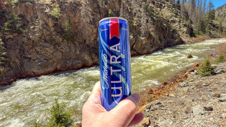 Hand holding a can of Michelob Ultra up to a landscape with rocks, a river, a hillside, and trees