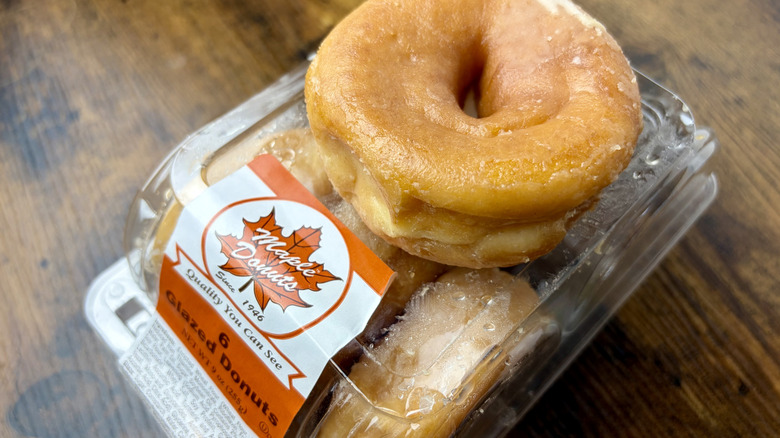A plastic container of Maple Donuts brand Glazed Donuts with one glazed donut on top of container