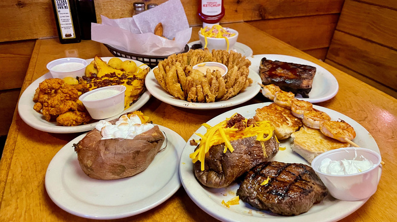 Multiple plates of food including steak, baked potato and Cactus Blossom on table at Texas Roadhouse rolls
