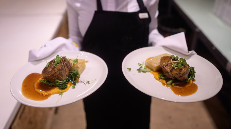 A server in white shirt and black apron carrying two steak dishes