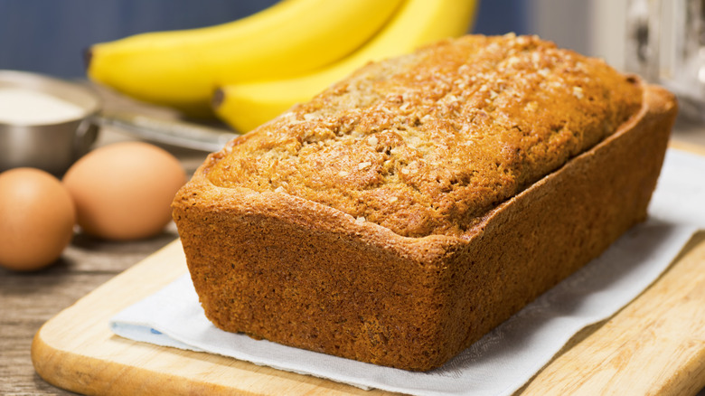 Banana bread with bananas, eggs, and measuring cup of sugar in the background