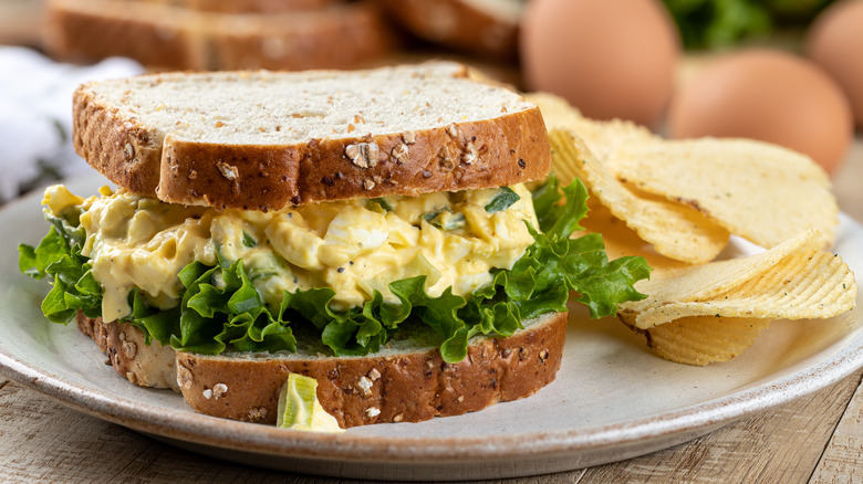 A close-up of an egg salad sandwich on wheat bread with potato chips on the side