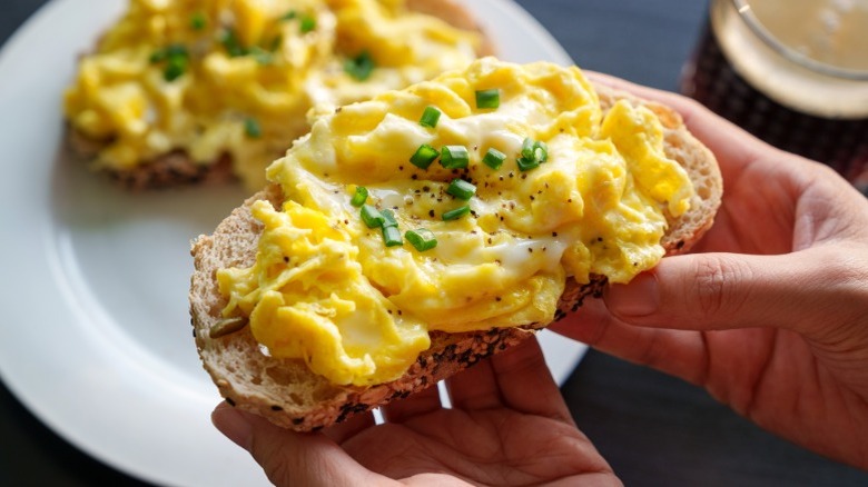Hands holding creamy scrambled eggs on toast, finished with black pepper and scallion.