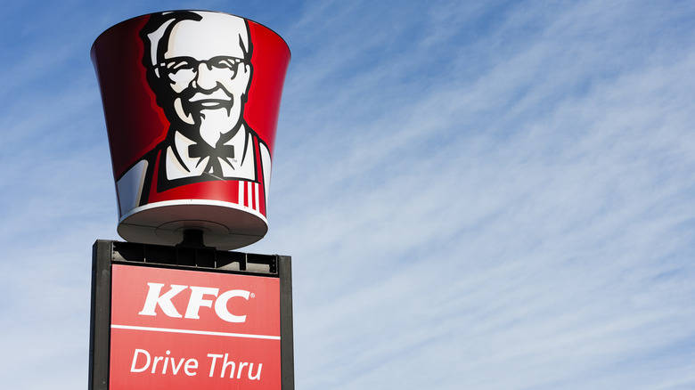 A KFC bucket with Colonel Sanders' image on top of a KFC Drive Thru sign against blue sky
