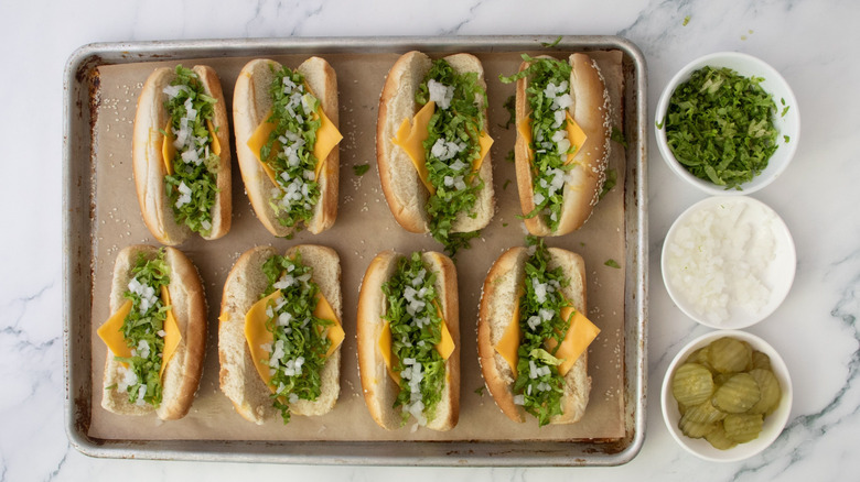 Hot dog buns filled with cheese, lettuce, and onion next to bowls of toppings