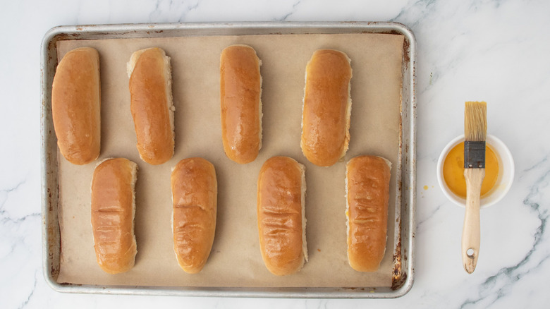 Hot dog buns on baking sheet next to egg wash and pastry brush