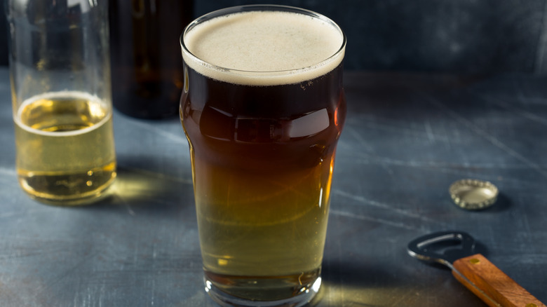 A dark beer and cider snakebite in a pint glass on a worn table near a bottle opener