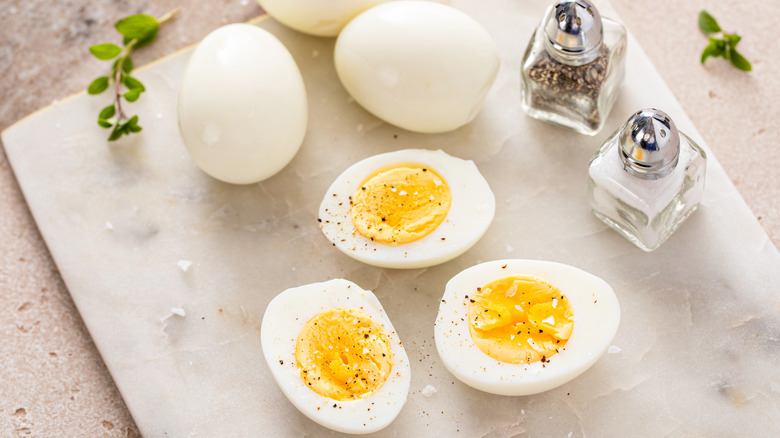 A stone cutting board with boiled eggs, some sliced in half with salt and pepper, some uncut but peeled.