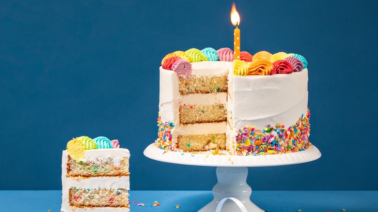 A funfetti birthday cake with a slice of cake beside it, covered in sprinkles and topped with a lit candle