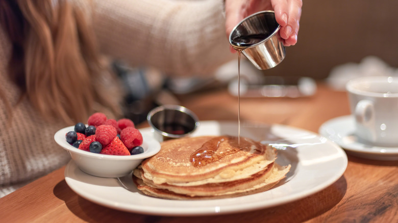 Woman pouring syrup on stack of pancakes with side of fresh berries at restaurant
