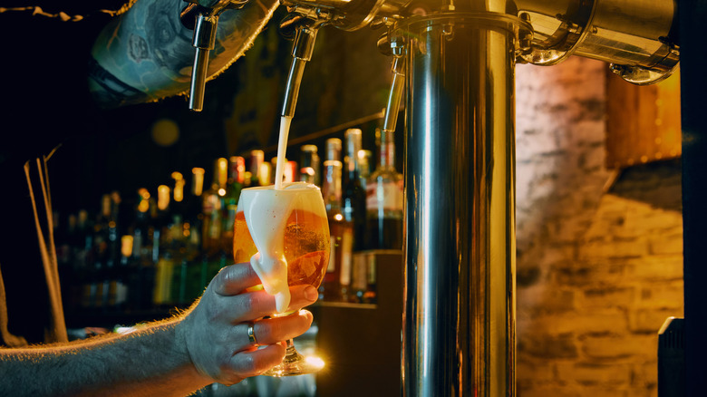 A hand fills a goblet beer glass with draft beer and foam overflowing over the top.