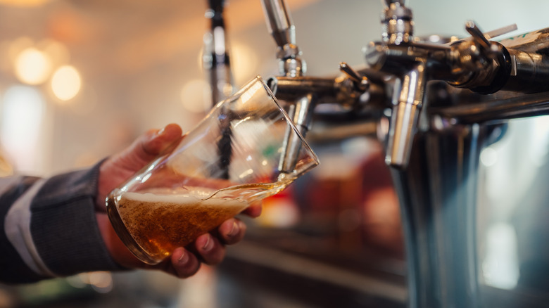 A hand fills a tilted pint glass from a beer tap with the nozzle dipped in