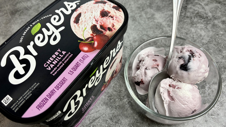 A carton of Breyers Cherry Vanilla ice cream next to a clear bowl with scoops of the ice cream and a spoon